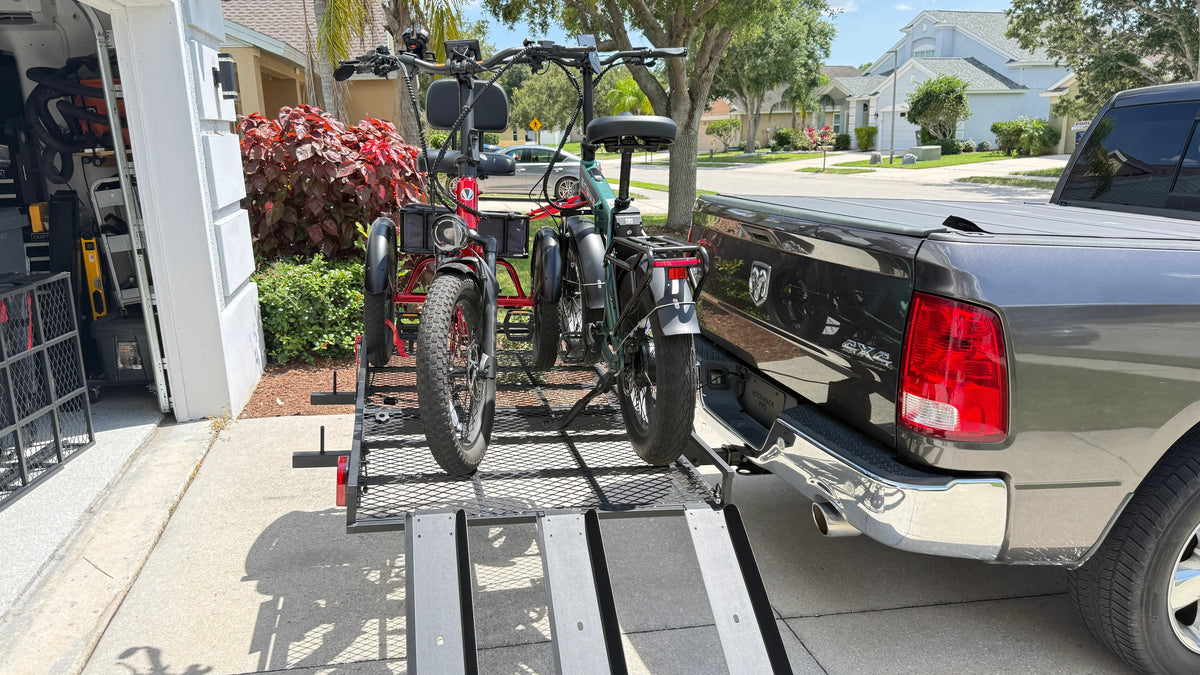 A black electric cargo bike and a red bicycle are mounted on a metal trailer hitch attached to the rear of a dark pickup truck, parked outside a residential garage with a street and trees in the background.