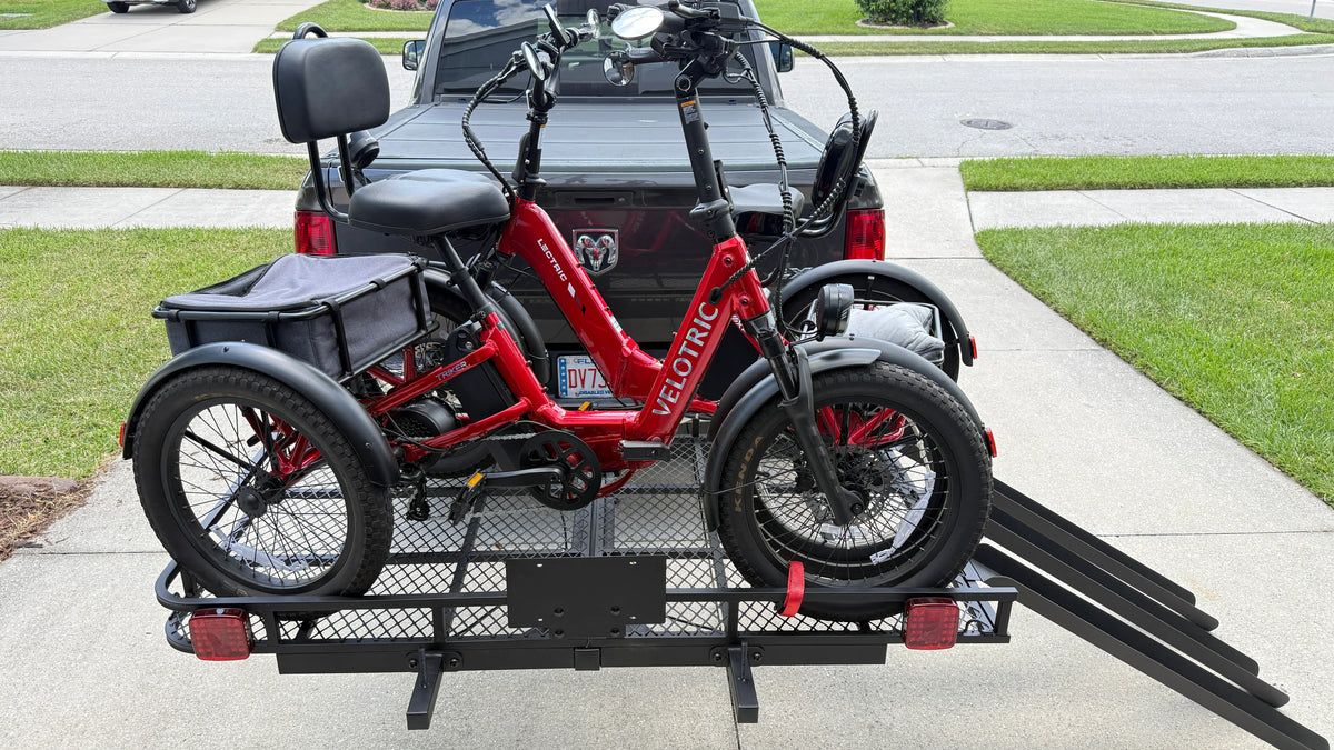 A red electric tricycle with a rear cargo basket is mounted on a black metal hitch carrier rack attached to the back of a black pickup truck, parked on a concrete driveway with grass and a street in the background.