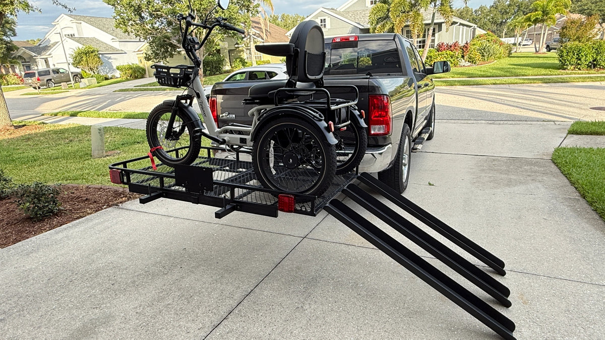 A black wheelchair cargo carrier hitched to the rear of a dark pickup truck, with a wheelchair mounted on the frame and a bicycle secured on the front rack, set on a concrete driveway in a suburban neighborhood.