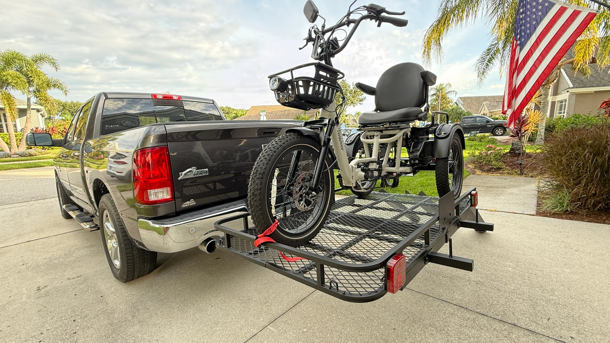 A black electric tricycle with a rear wheelchair seat is mounted on a black metal utility trailer hitched to the bed of a black pickup truck, parked on a concrete driveway in a residential neighborhood with an American flag in the background.